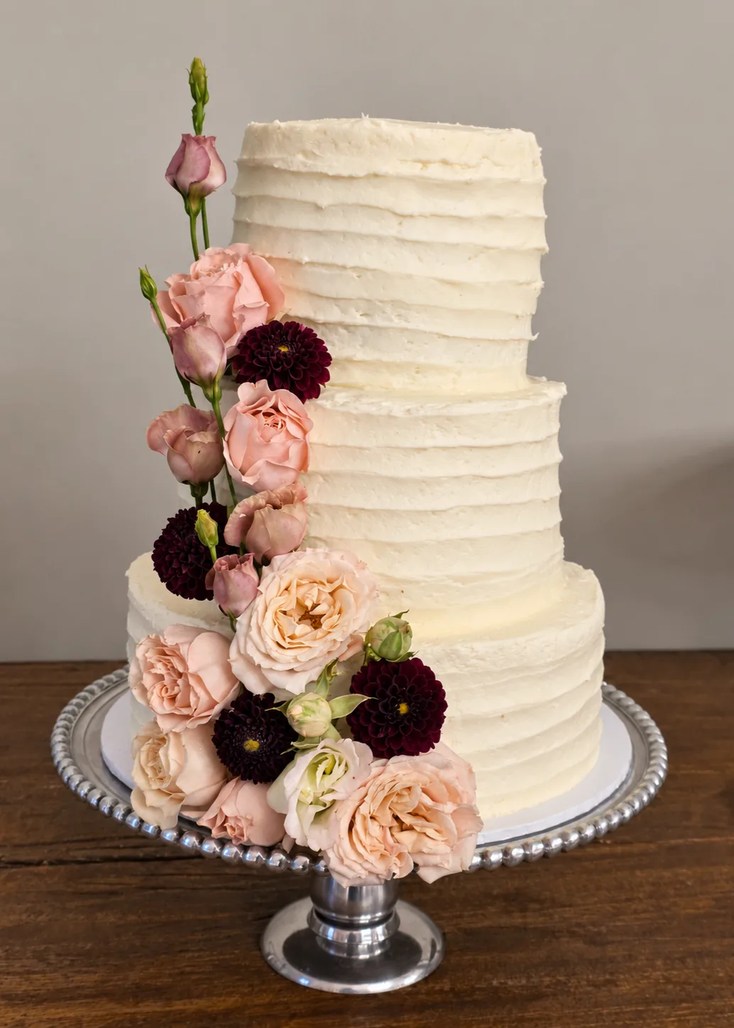 Three tier textured buttercream wedding cake with a cascade of blush roses and burgundy dahlias on a silver cake stand.
