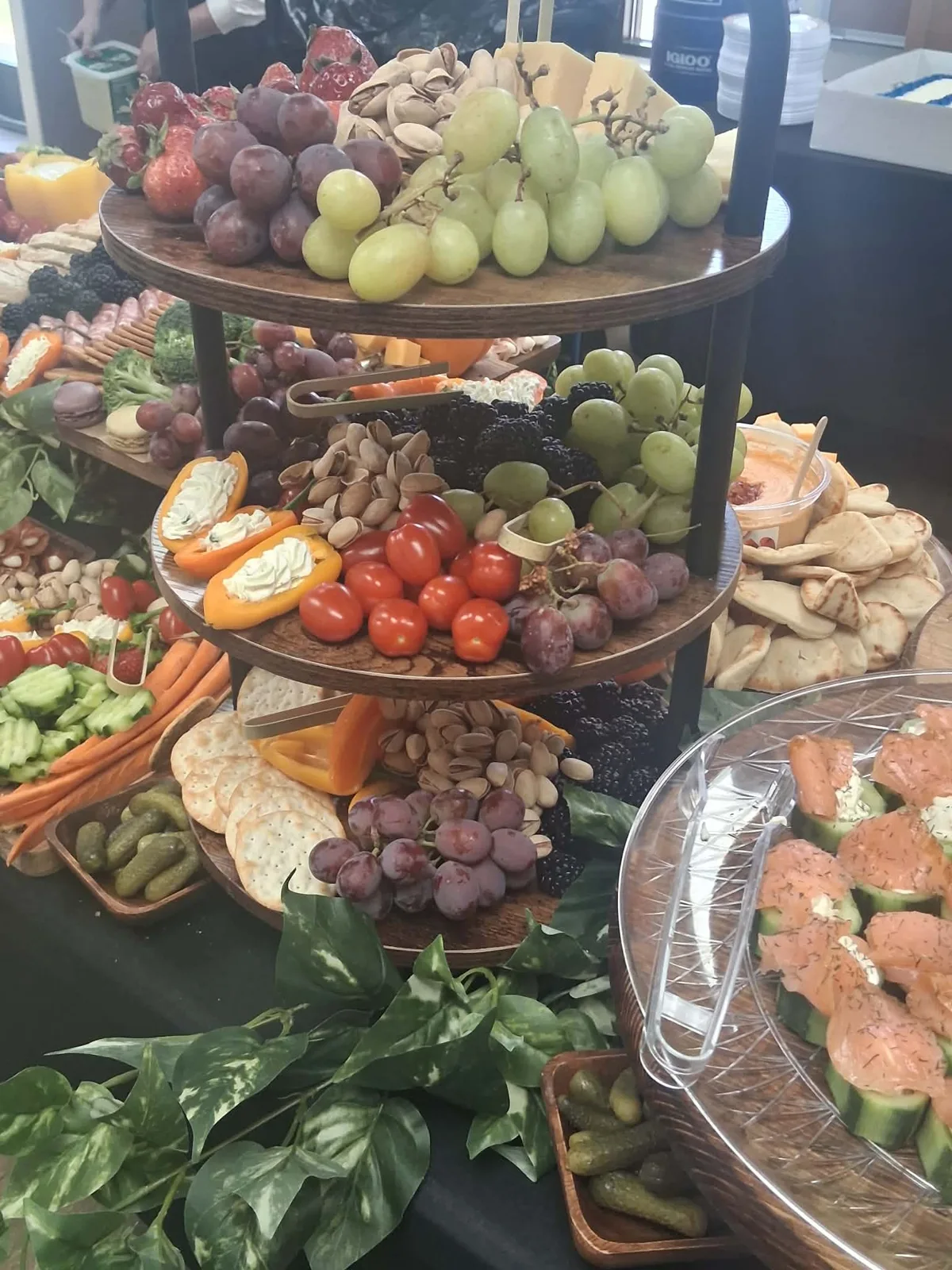 Three tier grazing stand with grapes, pistachios, tomatoes, stuffed mini peppers and crackers, beside a tray of smoked salmon cucumber bites.