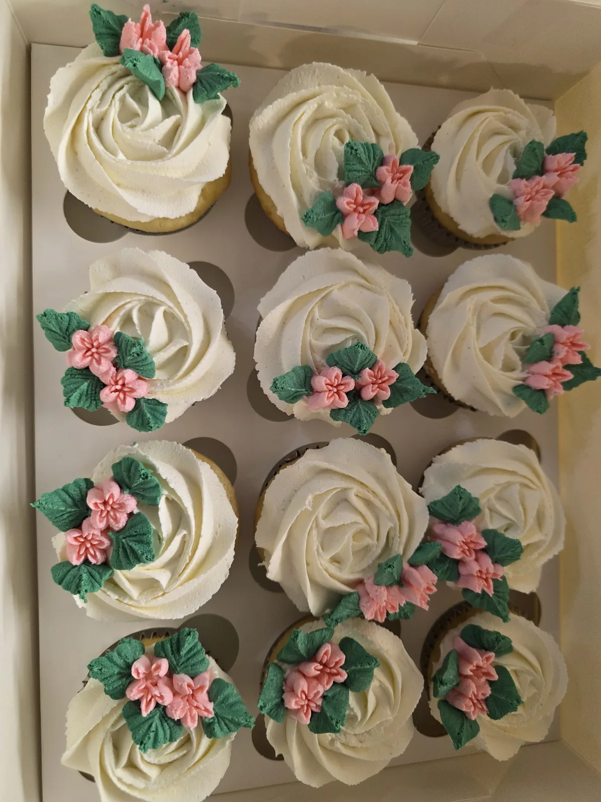 A box of a dozen vanilla rose swirl cupcakes topped with small pink buttercream flowers and green leaves.